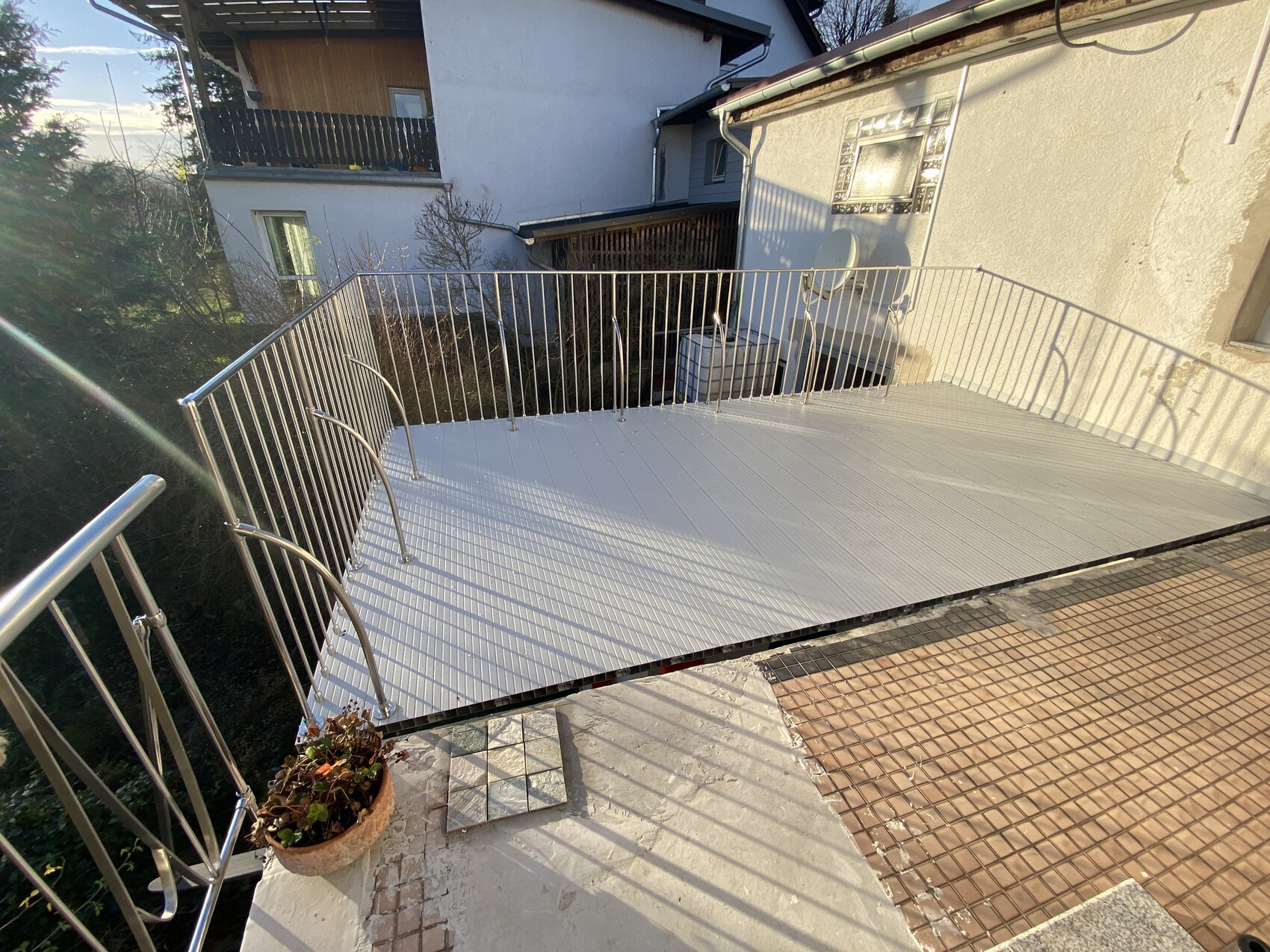 Heller Balkon mit Metallgeländer, weiße Bodenplatten, Topfpflanze; Nachbarhäuser im Hintergrund.