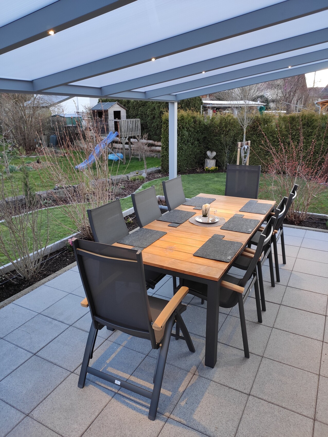 Gedeckter Holztisch mit Stühlen unter einer Überdachung; Garten mit Spielhaus im Hintergrund.