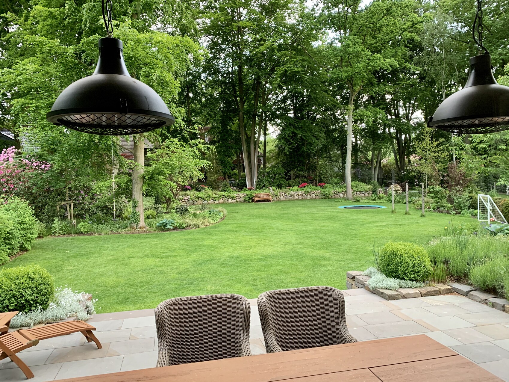 Gartenansicht: Terrasse mit Tisch und Korbstühle im Vordergrund, grüne Wiese, Bäume und Beete.