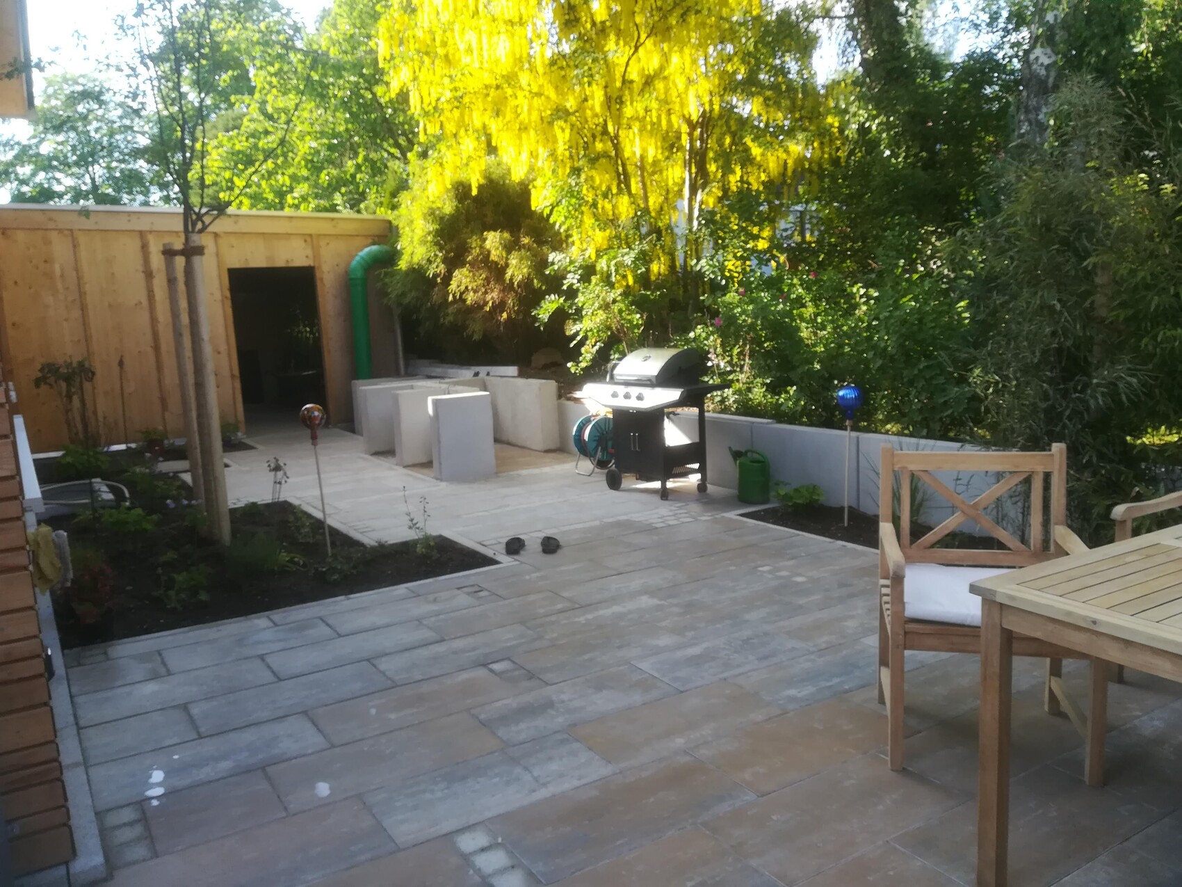 Garten-Terrasse mit Holztisch und Stühlen, Grill, Pflanzbeeten und grüner Hecke im Hintergrund.
