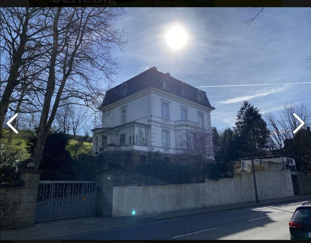 Großes weißes Villahaus auf einer Anhöhe, umgeben von Mauer und Bäumen, sonniger Himmel.
