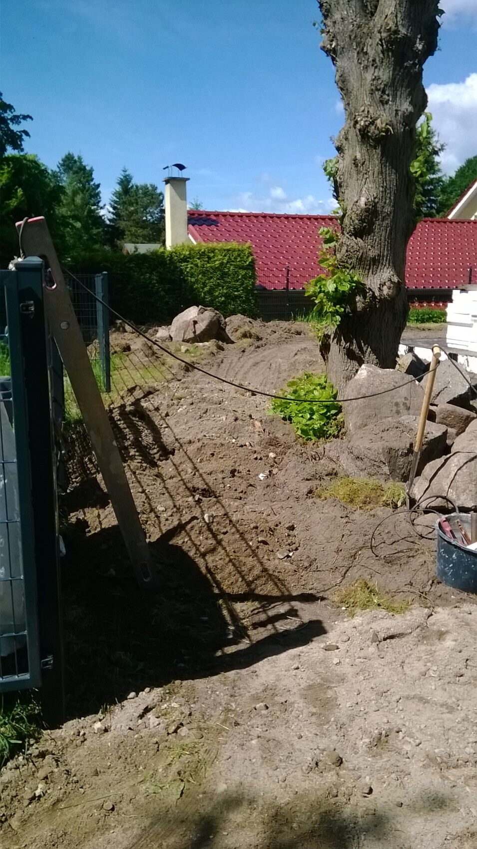 Gartenbaustelle mit Baum, Erdarbeiten, Zaun, Leiter und Steinen; rotes Dach im Hintergrund.