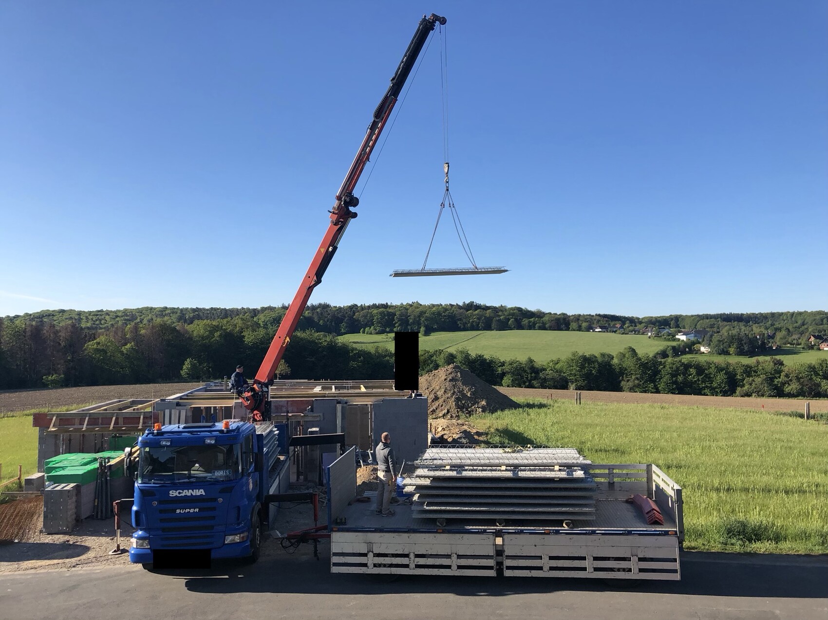 Kran hebt lange Stahlplatte über Baugrube; blauer Scania-Lkw, Bauarbeiter, Metallplatten.