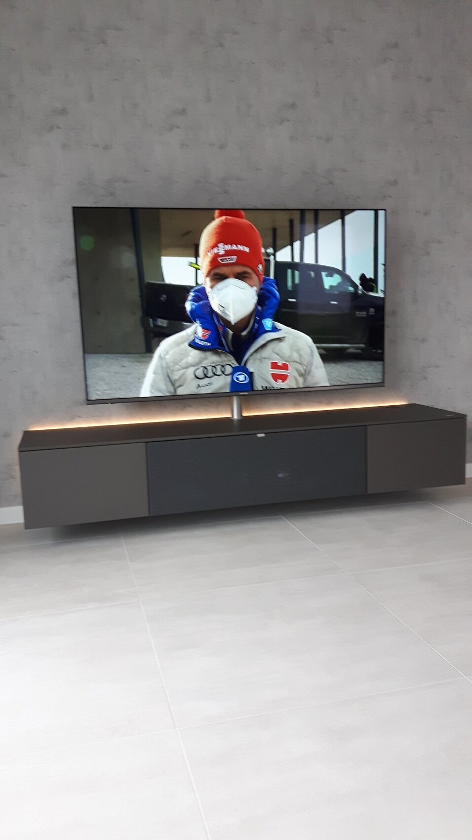 TV zeigt Mann mit weißer Maske, roter Mütze und blauer Jacke, Auto im Hintergrund