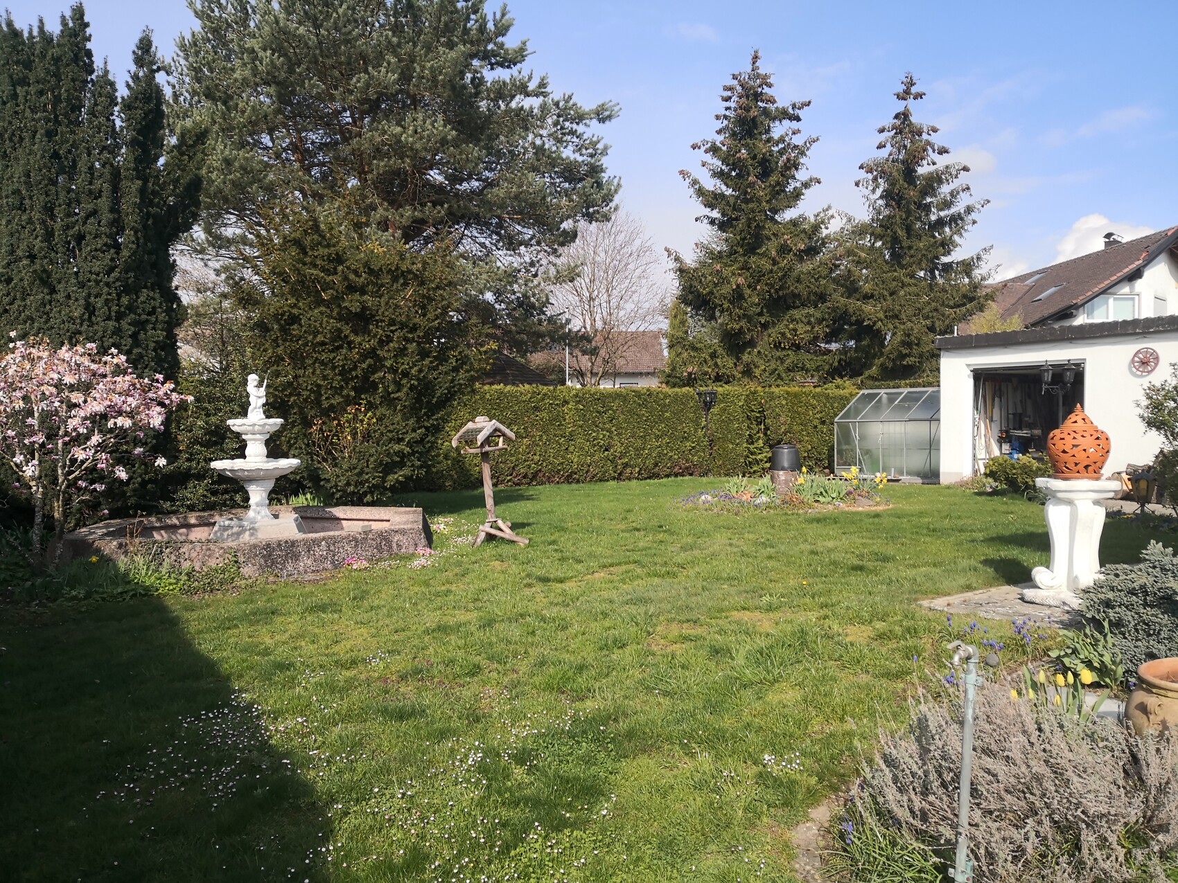 Garten mit Brunnenstatue, grünem Rasen, Gewächshaus und orange Zierkugel auf Sockel.
