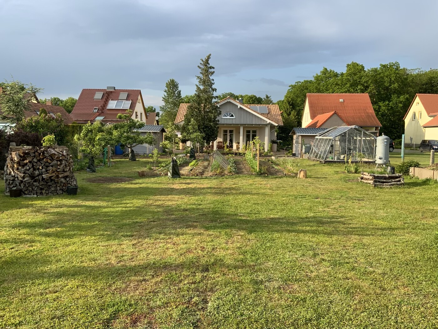 Garten mit Gemüsebeeten, Glasdach-Gewächshaus, Holzstapel links, Einfamilienhaus im Hintergrund.