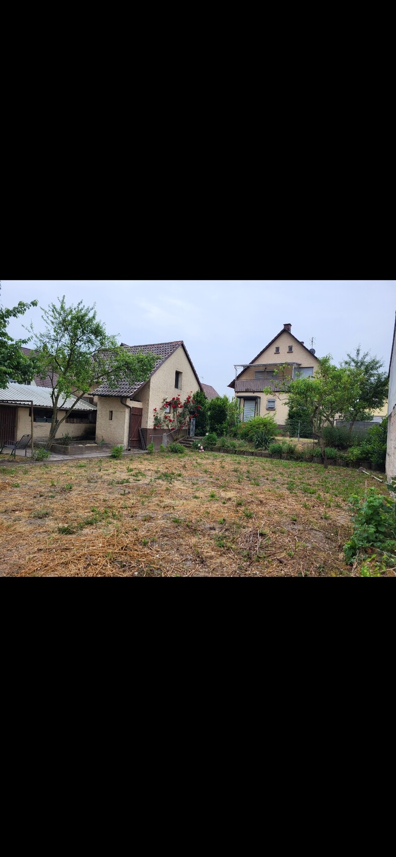 Zwei beige Häuser umgeben einen Garten mit Rosen, Bäumen und trockenem Boden.