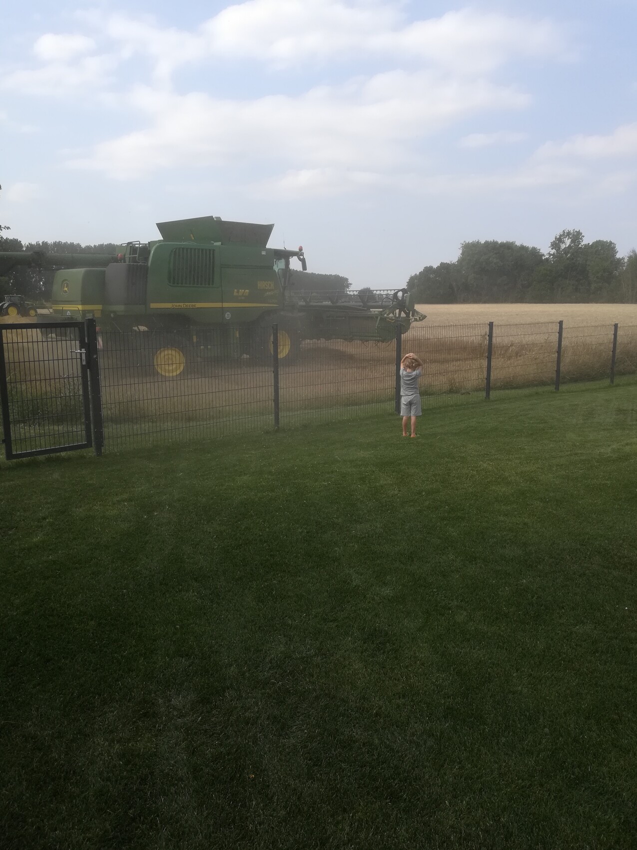 Kleines Kind vor Zaun beobachtet großen grünen Mähdrescher auf staubigem Feld.