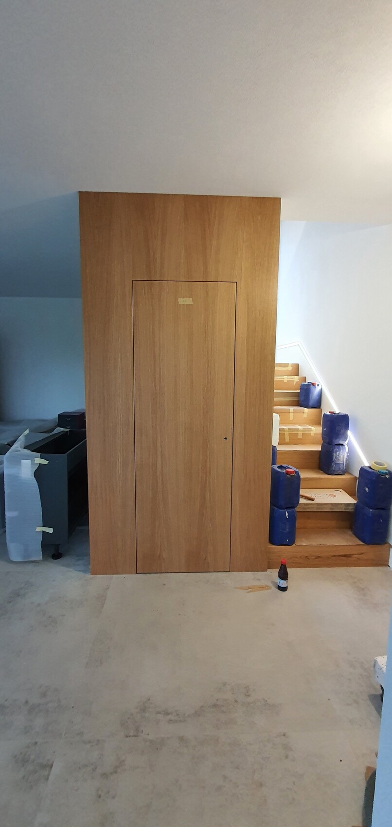 Großer brauner Holzschrank im Keller, rechts Treppe mit blauen Kanistern; Flasche davor.