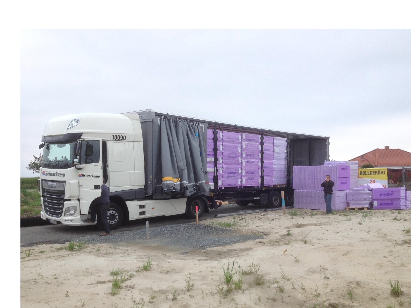 Weißer Lastwagen mit offenem Trailer transportiert violette Dämmplatten auf einer Baustelle.