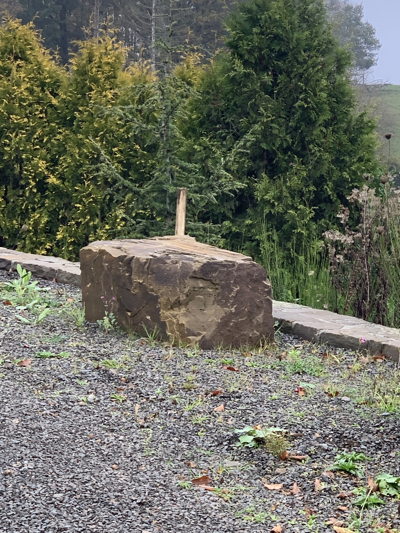 Großer Steinblock mit Holzstab darauf, Kiesboden und grüne Hecke dahinter
