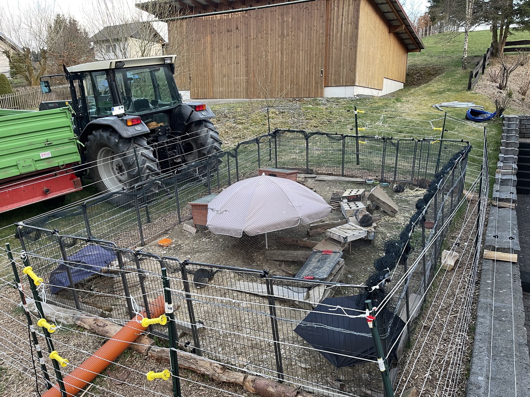 Garten-Gehege mit Metallzaun, Traktor links, runder Schirm und Baumholz im Hof.