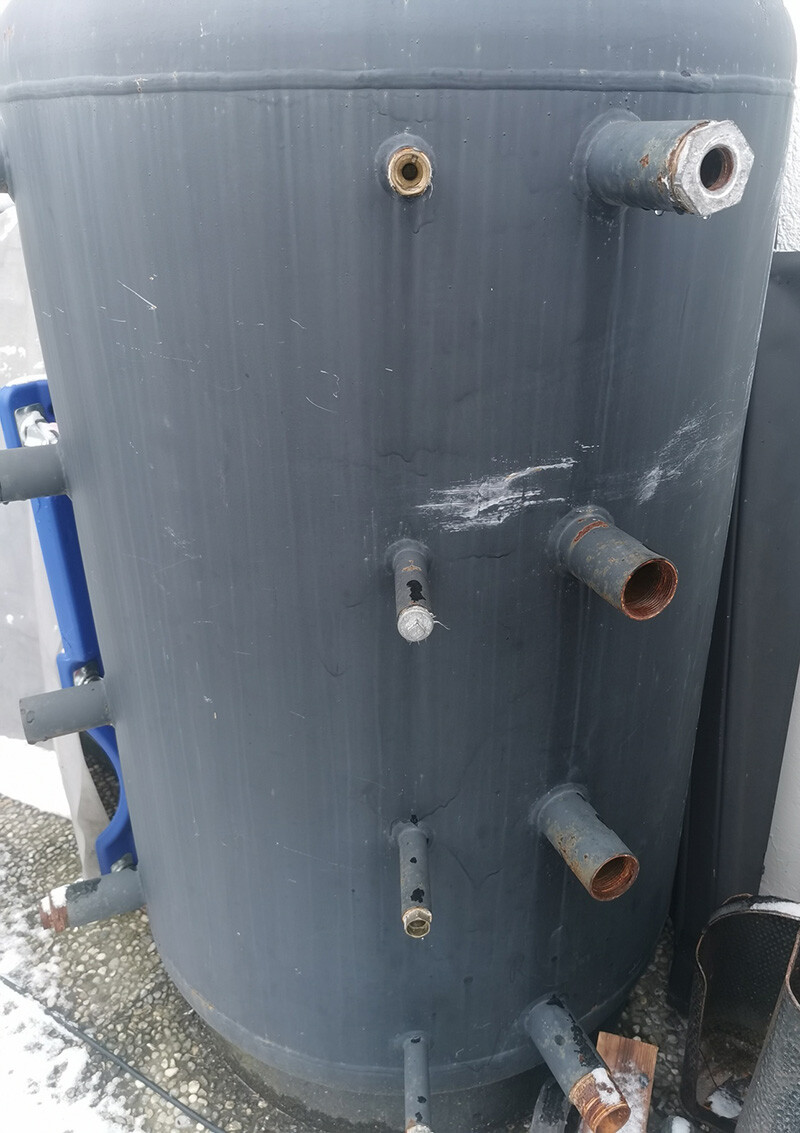 Grauer vertikaler Wassertank im Freien mit mehreren rostigen Außenrohranschlüssen.