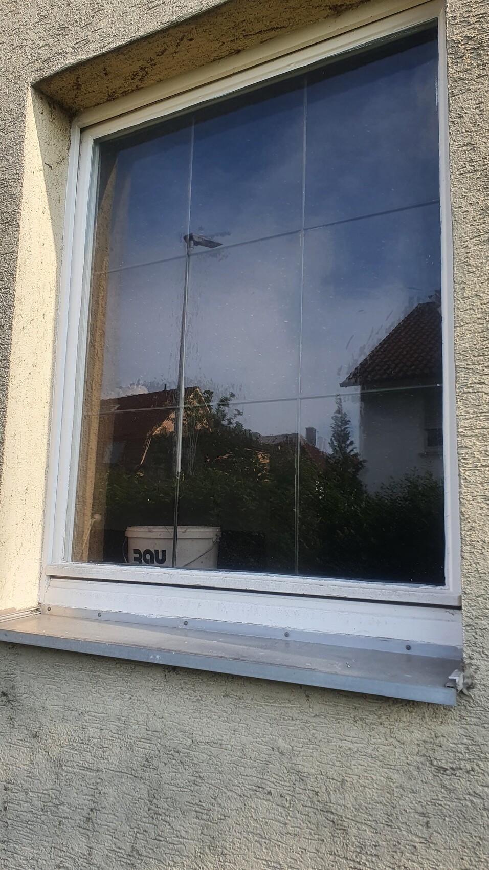 Fenster eines beigefarbenen Hauses mit weißem Rahmen; Reflektionen von Himmel und Bäumen.