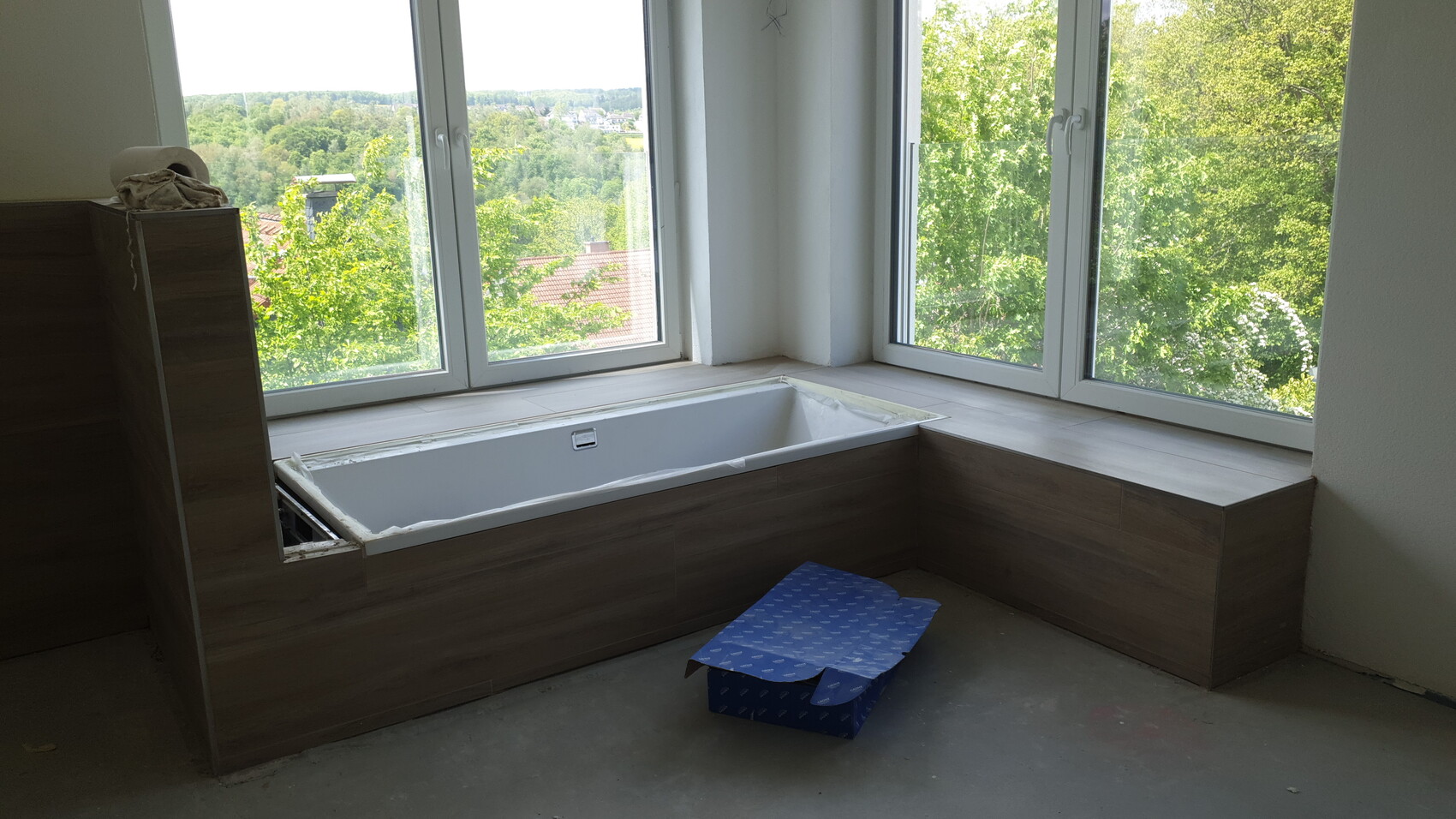 Badewanne in Holzverkleidung in Fensternische, Blick nach draußen; blaue Box auf dem Boden.