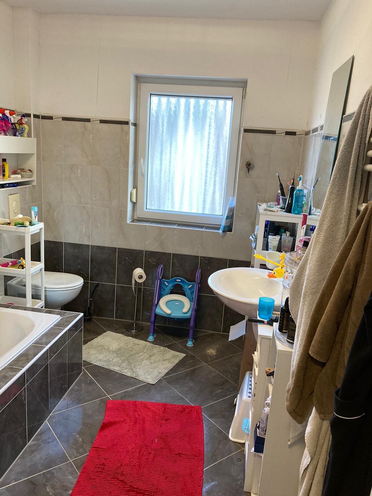 Badezimmer mit Waschbecken, Toilette, Badewanne, Fenster und Ablagen in grauen Fliesen.