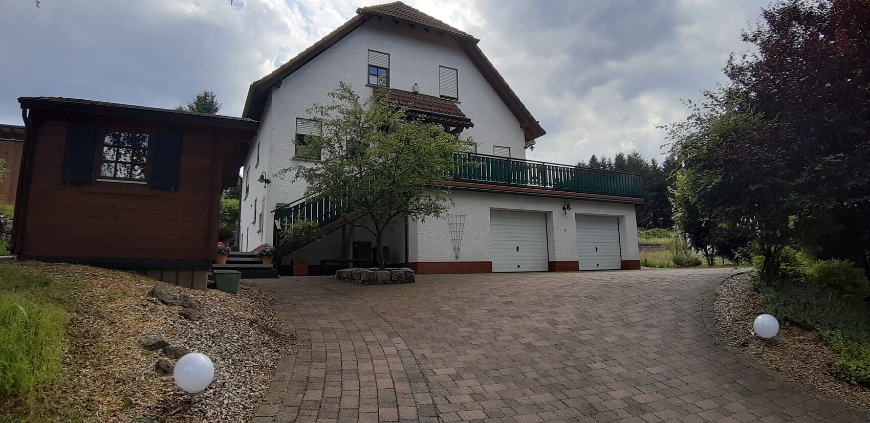 Weißes zweistöckiges Haus mit grünem Balkon, zwei Garagen, gepflasterter Hof und Baum davor.