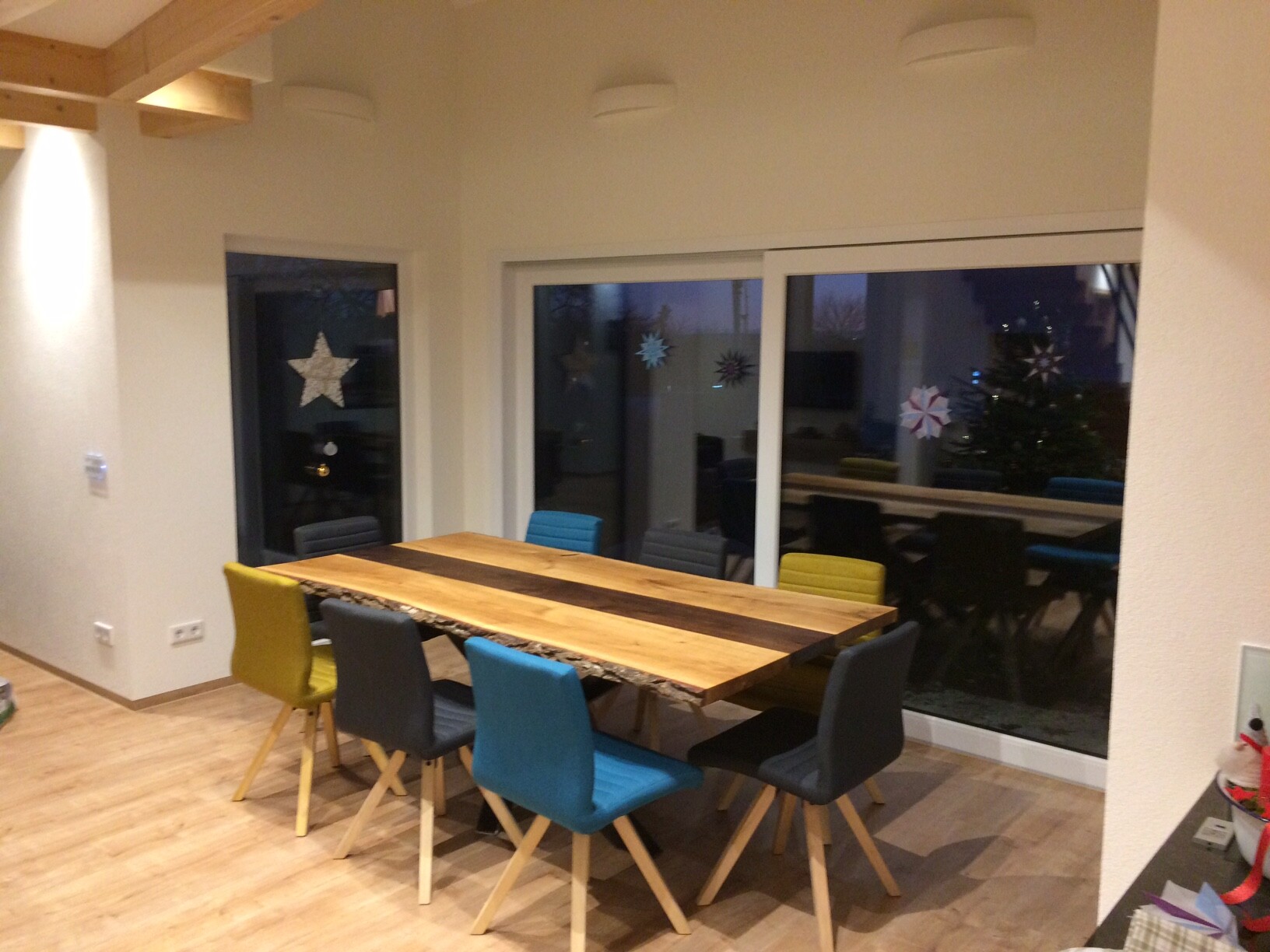 Großer Esstisch aus Holz mit bunten Stühlen in hellem Raum; Fenster mit Schneeflocken-Deko.
