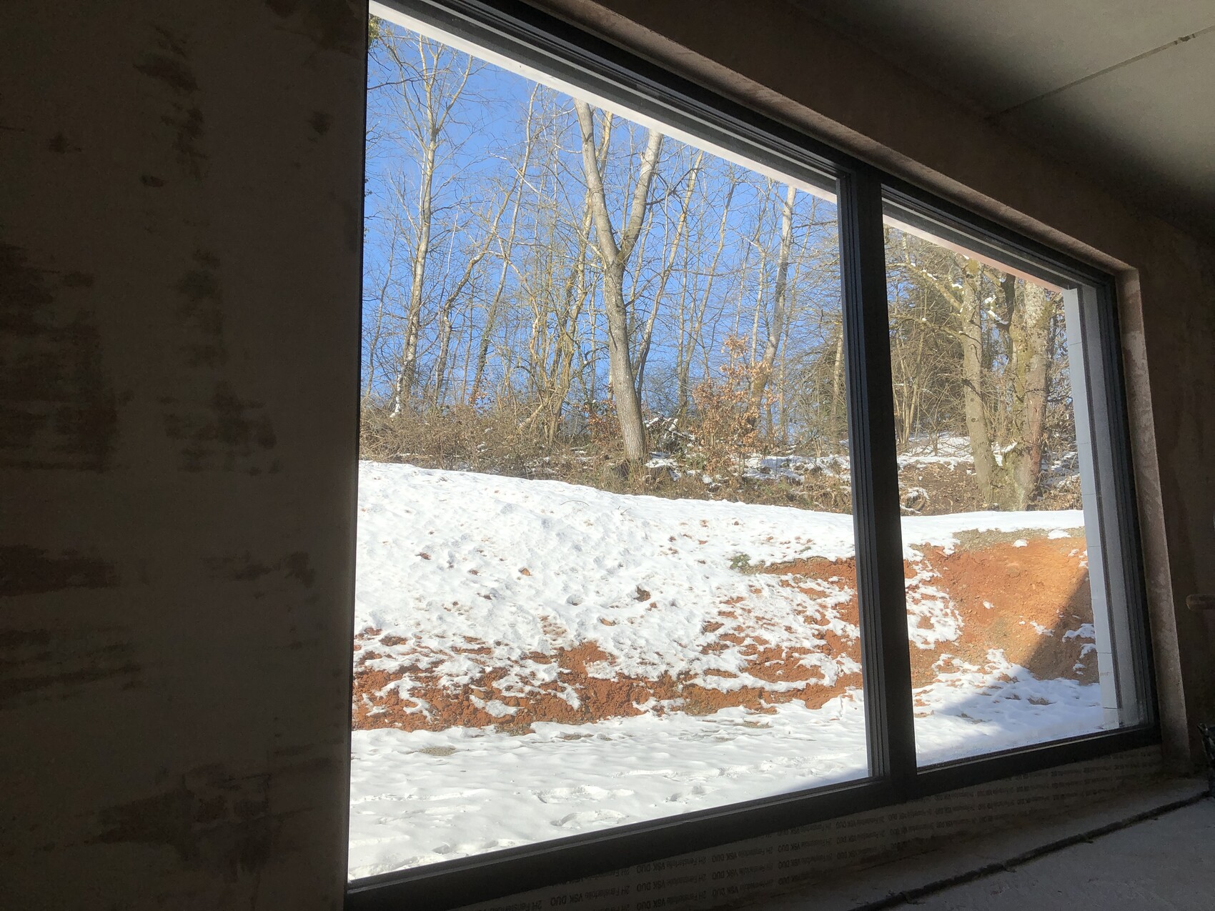 Blick durch großes Innenraumfenster auf verschneiten Boden, Erdwall und kahle Bäume im Freien