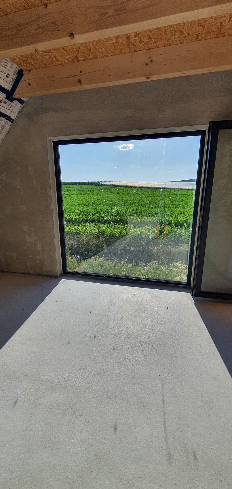 Große Fensterfront in einem unfertigen Innenraum; Blick auf grüne Felder und blauen Himmel.