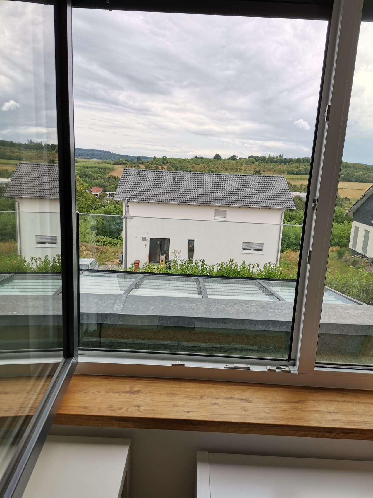 Offenes Fenster mit Fensterbank: Blick auf ein weißes Haus mit grauem Dach und Felder dahinter.