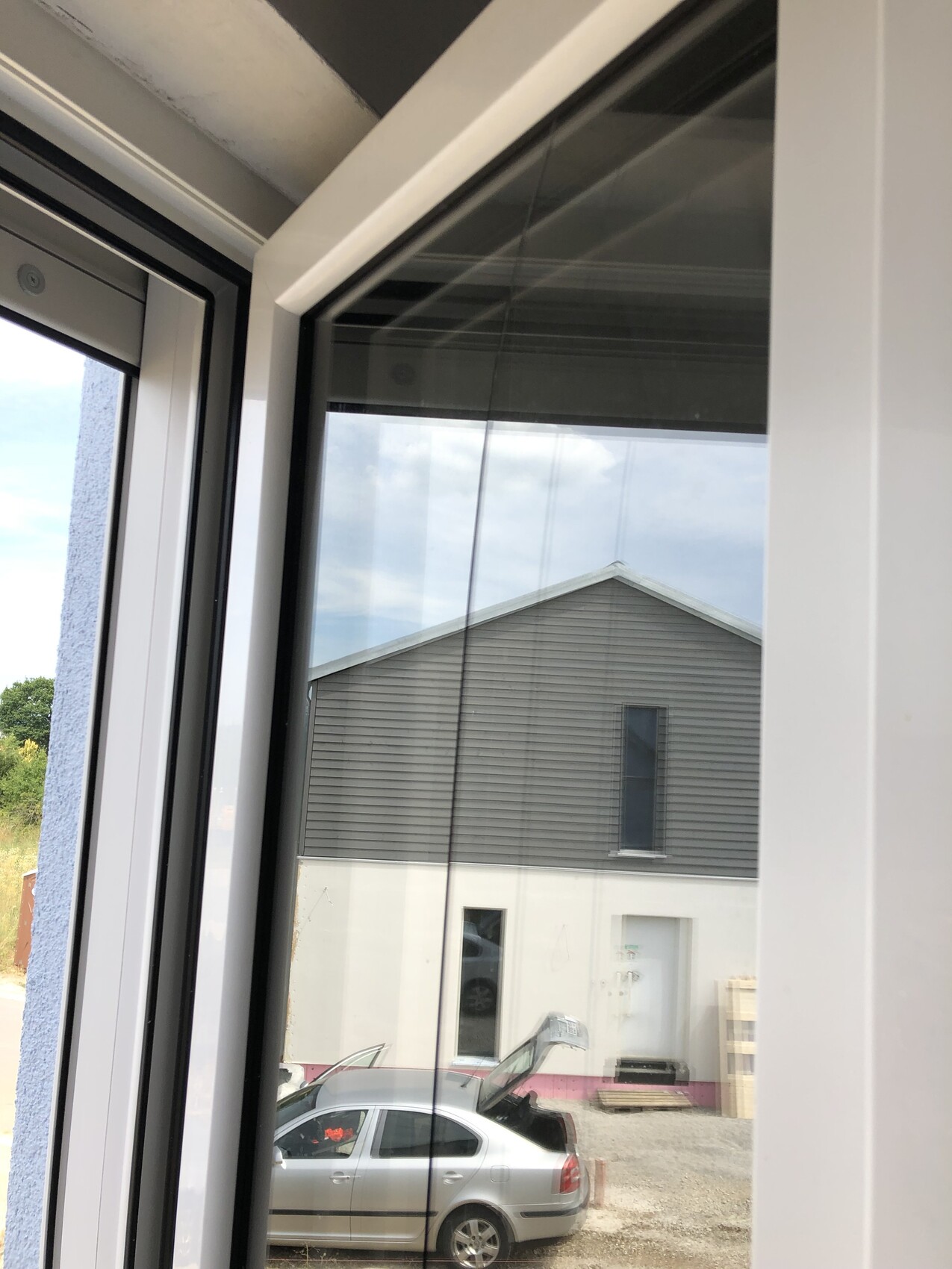 Blick durch Fenster auf graues Gebäude mit dunkler Fassade, Tür unten, silbernes Auto davor