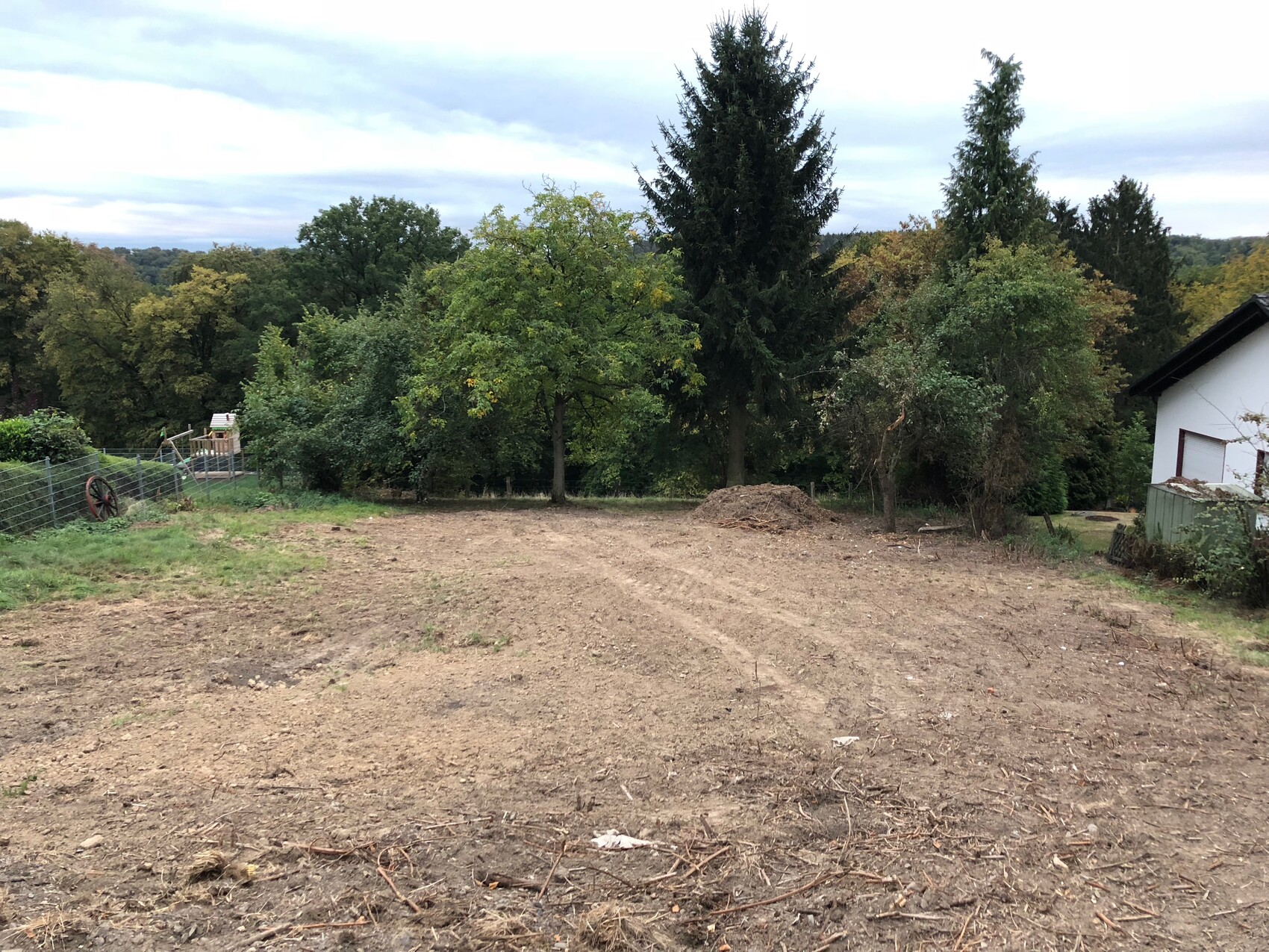 Freies Baugrundstueck mit Baeumen im Hintergrund, linker Zaun, altes Rad, rechts weisses Haus