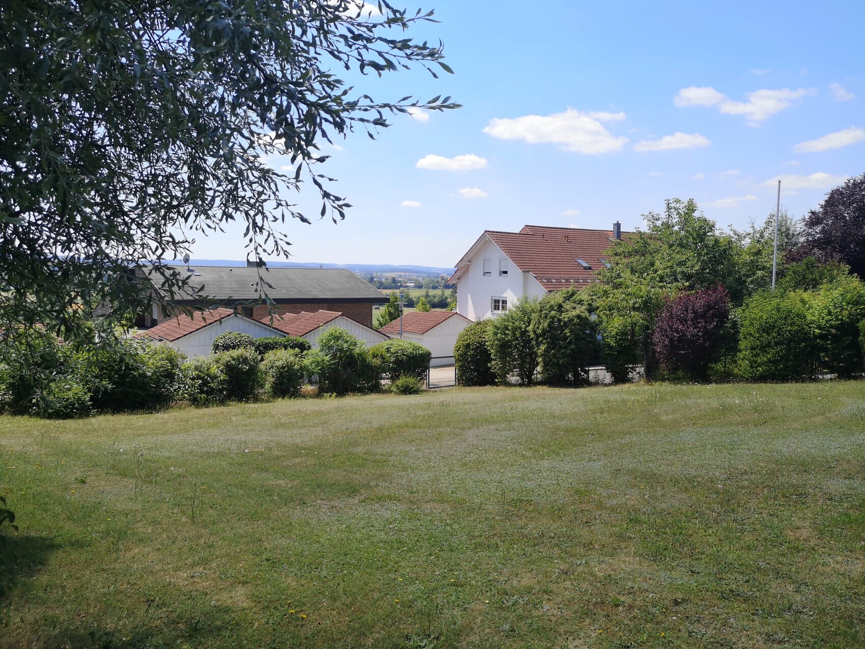 Grünes Grundstück mit Haus, Bäumen und Hecken unter blauem Himmel