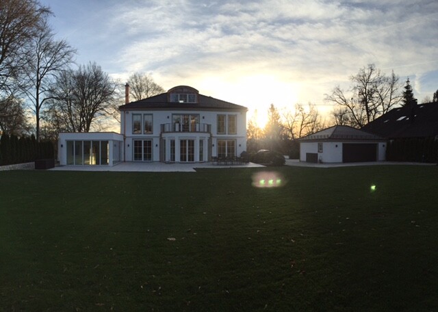 Großes weißes Haus mit Glasfront, Balkon und grünem Rasen bei Sonnenuntergang.