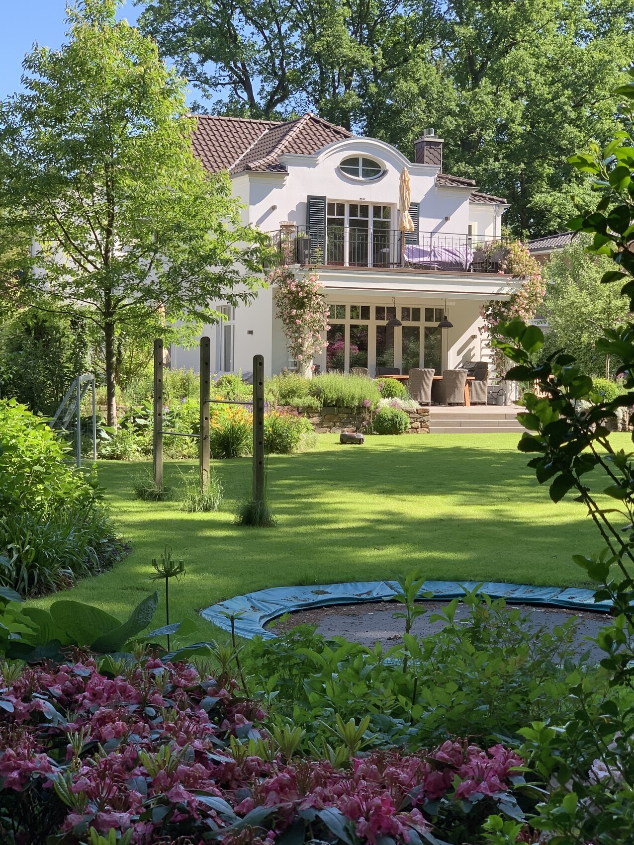 Weißes zweistöckiges Haus mit Balkon, Terrasse und Garten; Trampolin im Vordergrund und Blumenbeete.
