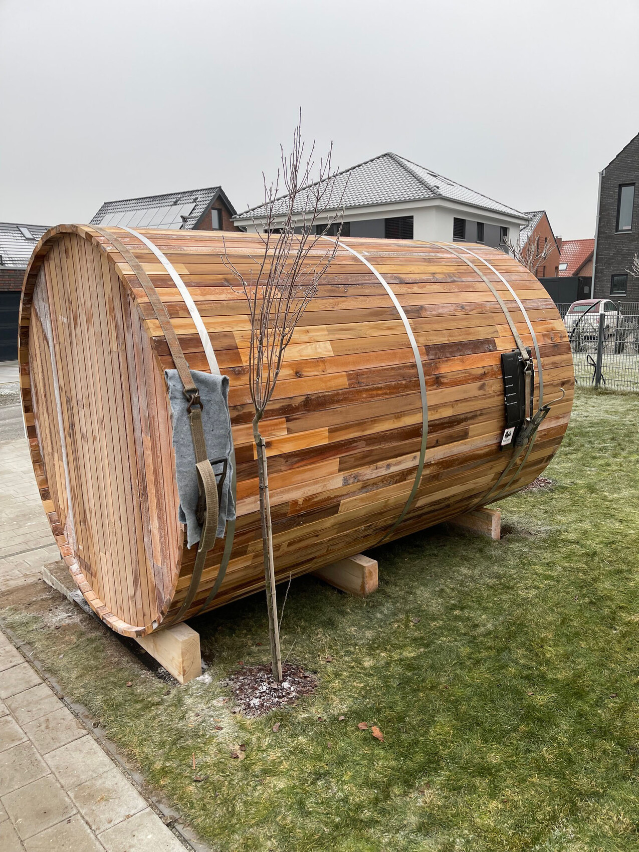Großes horizontales Holzfass mit Metallbändern auf Stützen im Garten, daneben ein Baum.