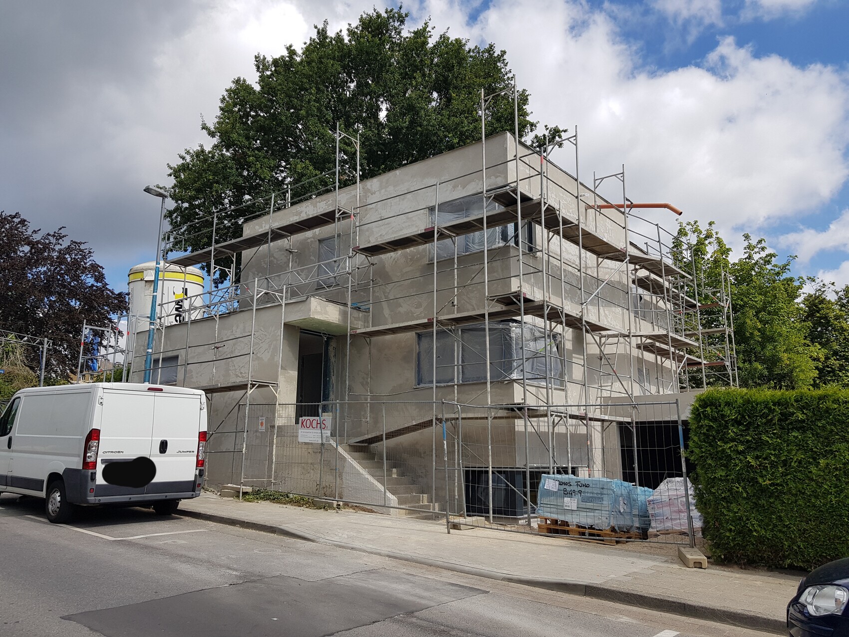 Gebäude unter Baugerüst mit Baustoffen und weißem Transporter davor; umliegende Bäume.
