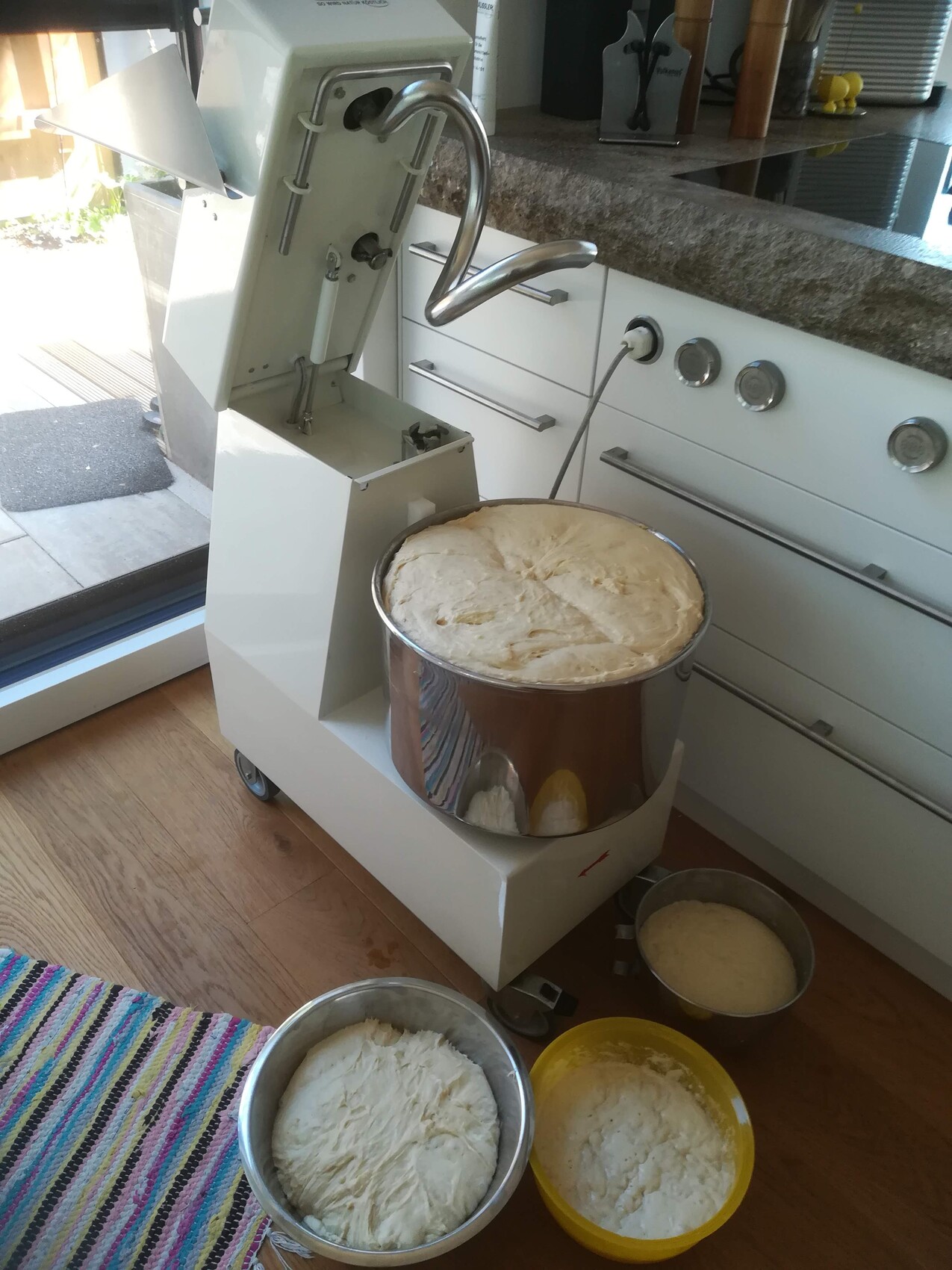 Großer weißer Küchenmixer mit offenem Deckel; daneben mehrere Teigschüsseln mit Hefeteig.