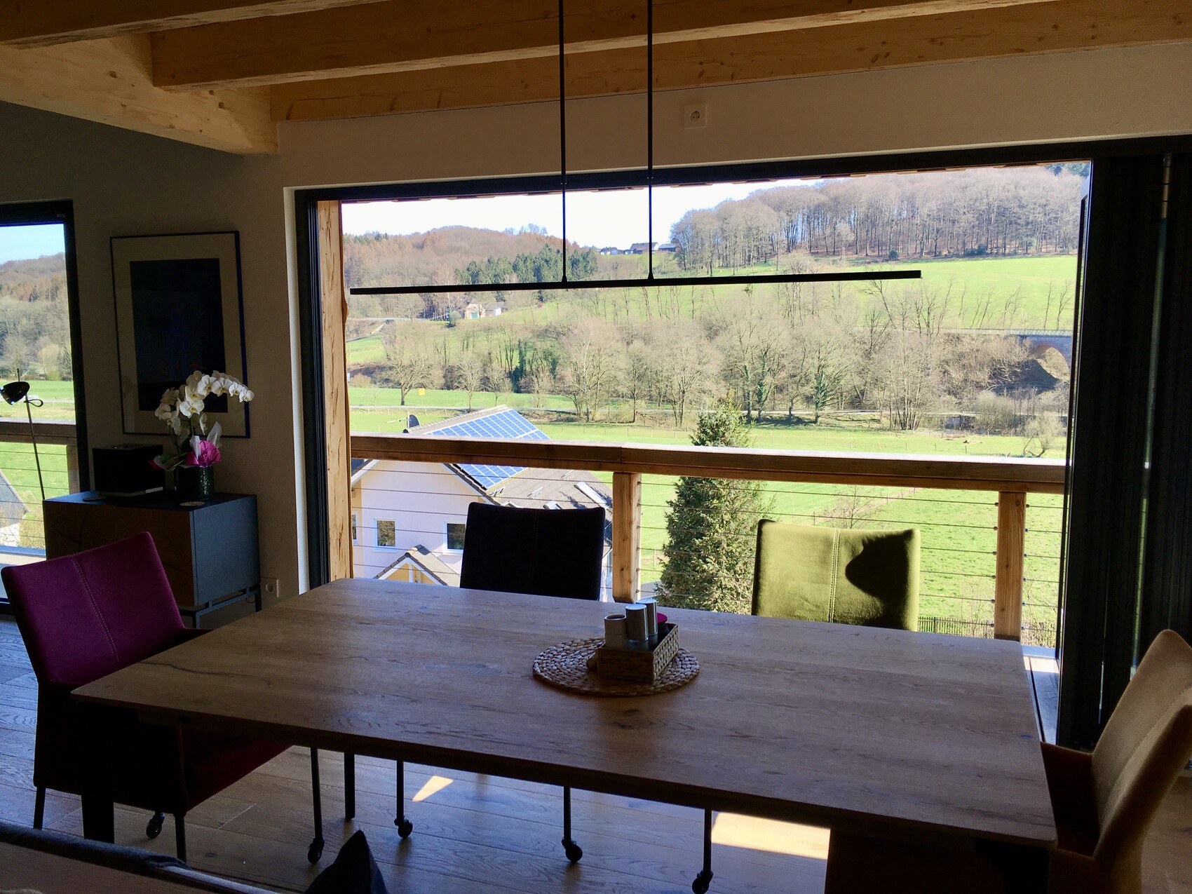 Esszimmer mit großer Fensterfront, Blick auf grüne Wiesen und Hügel; Holztisch und bunte Stühle.