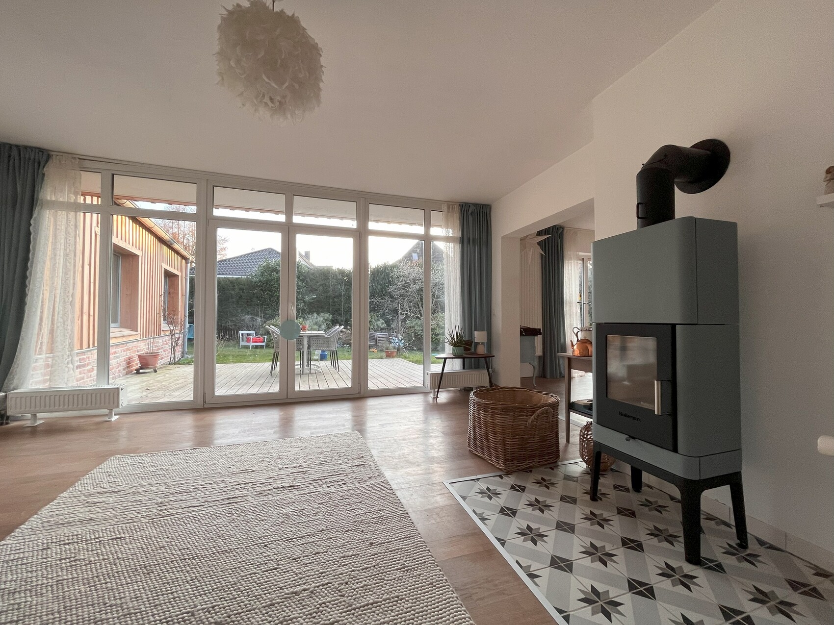 Wohnzimmer mit Holzofen rechts, große Glasfront zum Garten, Holzboden und Teppich.