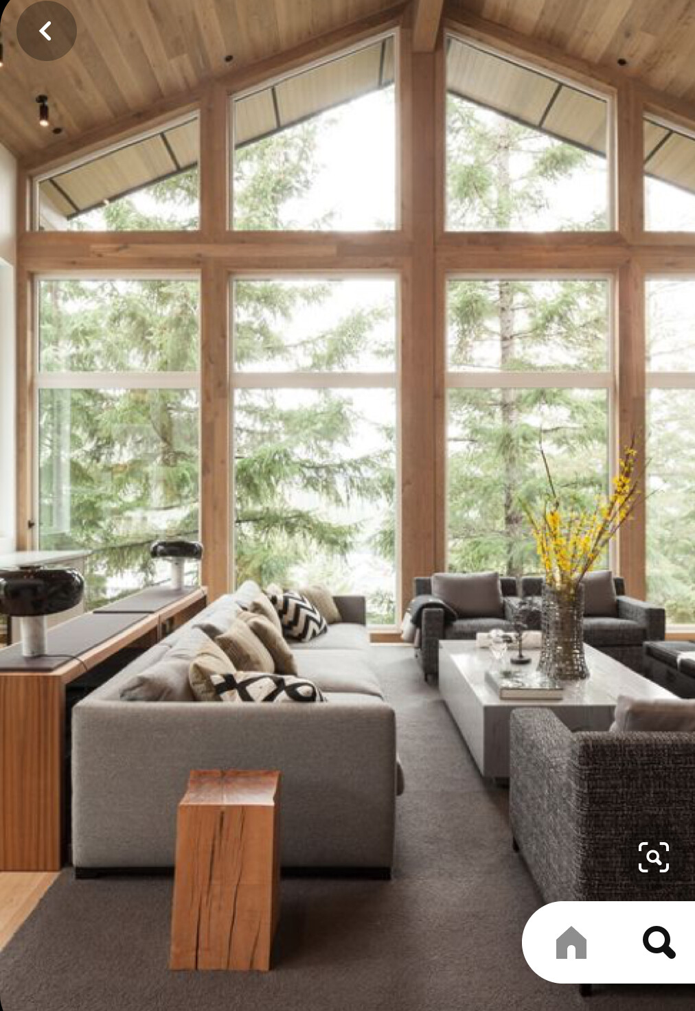 Helles Wohnzimmer mit großen Fensterfronten, Holzbalken, graues Sofa und Kaffeetisch.