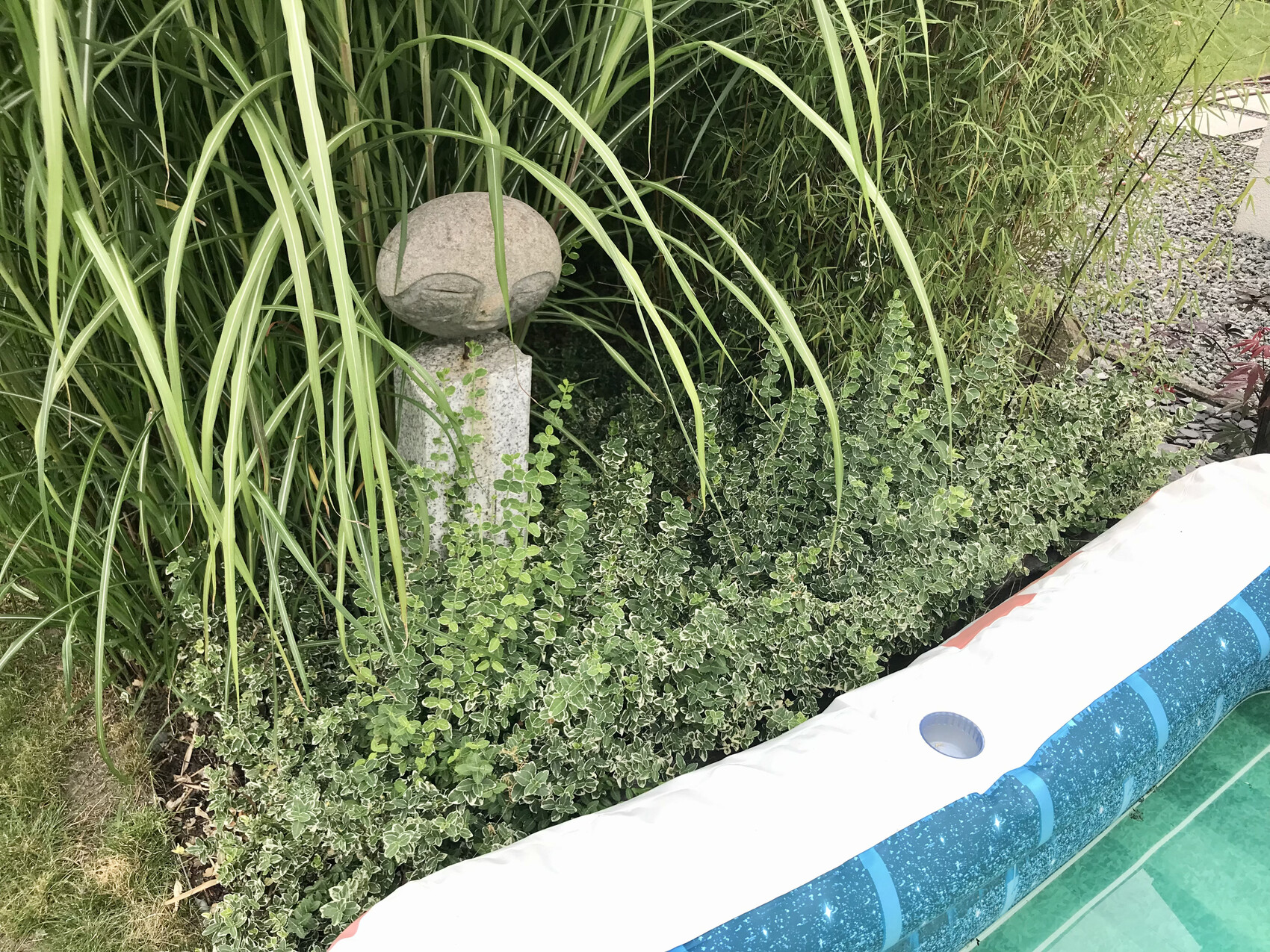 Steinkugel auf Sockel im Garten, umgeben von hohen Gräsern und grünem Bodendecker.