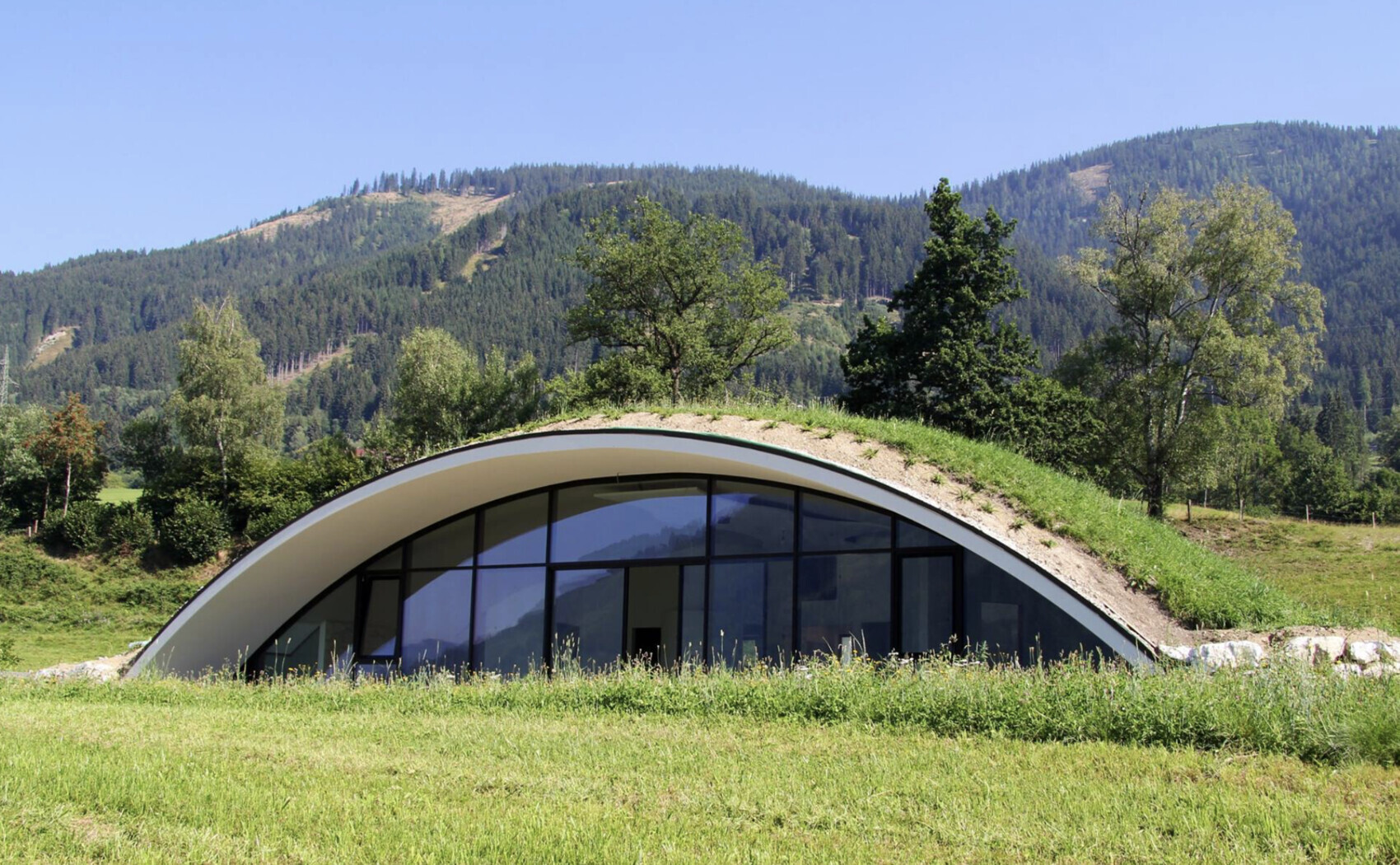 Gewölbtes Glasfront-Gebäude mit Grasdach, eingebettet in Wiesen vor Bergen