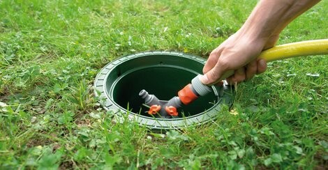 Person öffnet einen runden Gruben- bzw. Kanaldeckel im Gras, hält Werkzeug in der Hand.