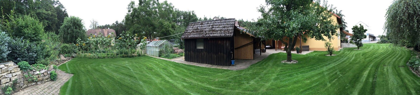 Gepflegter Garten mit grünem Rasen, Holzschuppen, Gewächshaus und Bäumen rund um ein Haus