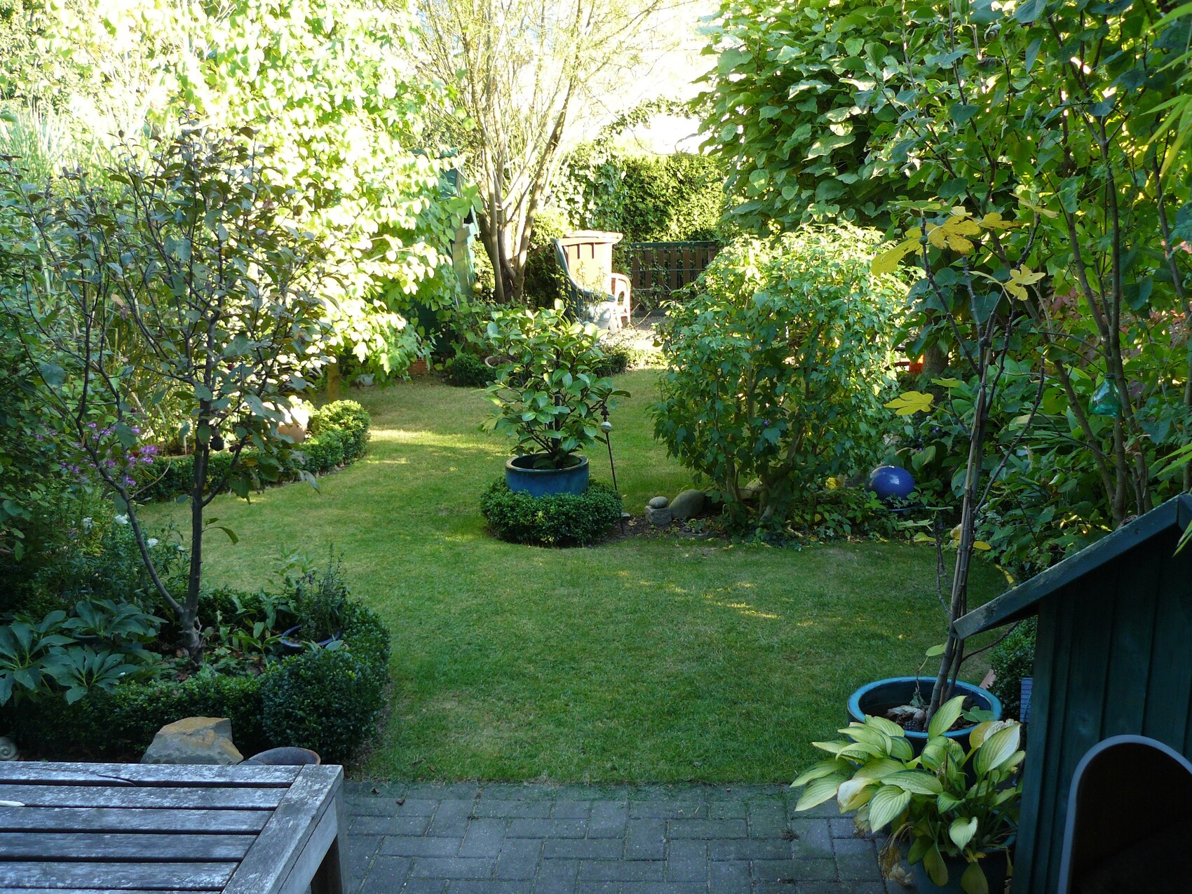 Garten mit grünem Rasen, Bäumen, Sträuchern, Topfpflanzen, blauem Ball und Sitzplatz im Hintergrund