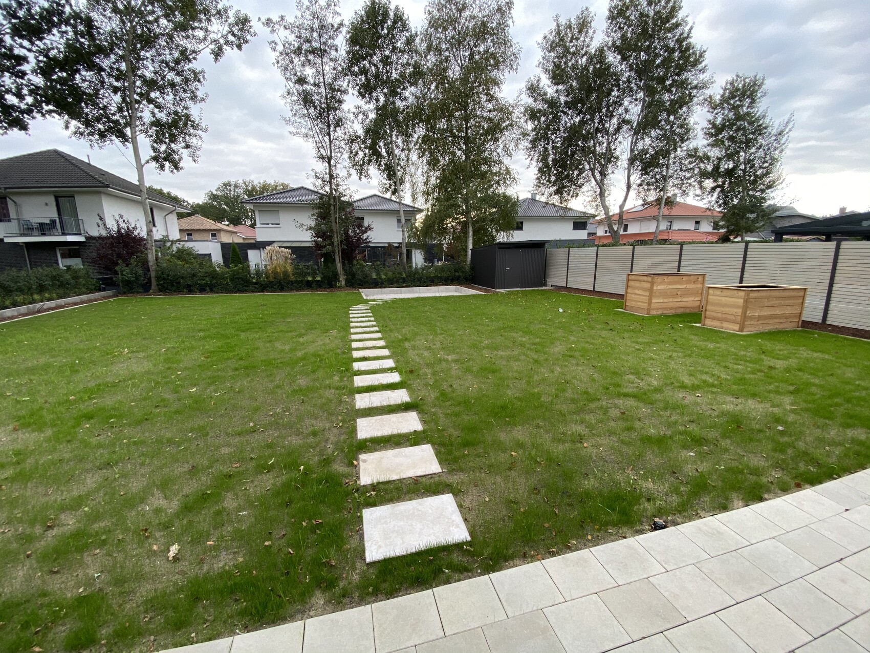 Garten mit Steinpfad über grünem Rasen, Holzplanter am Zaun, Wohnhäuser im Hintergrund.