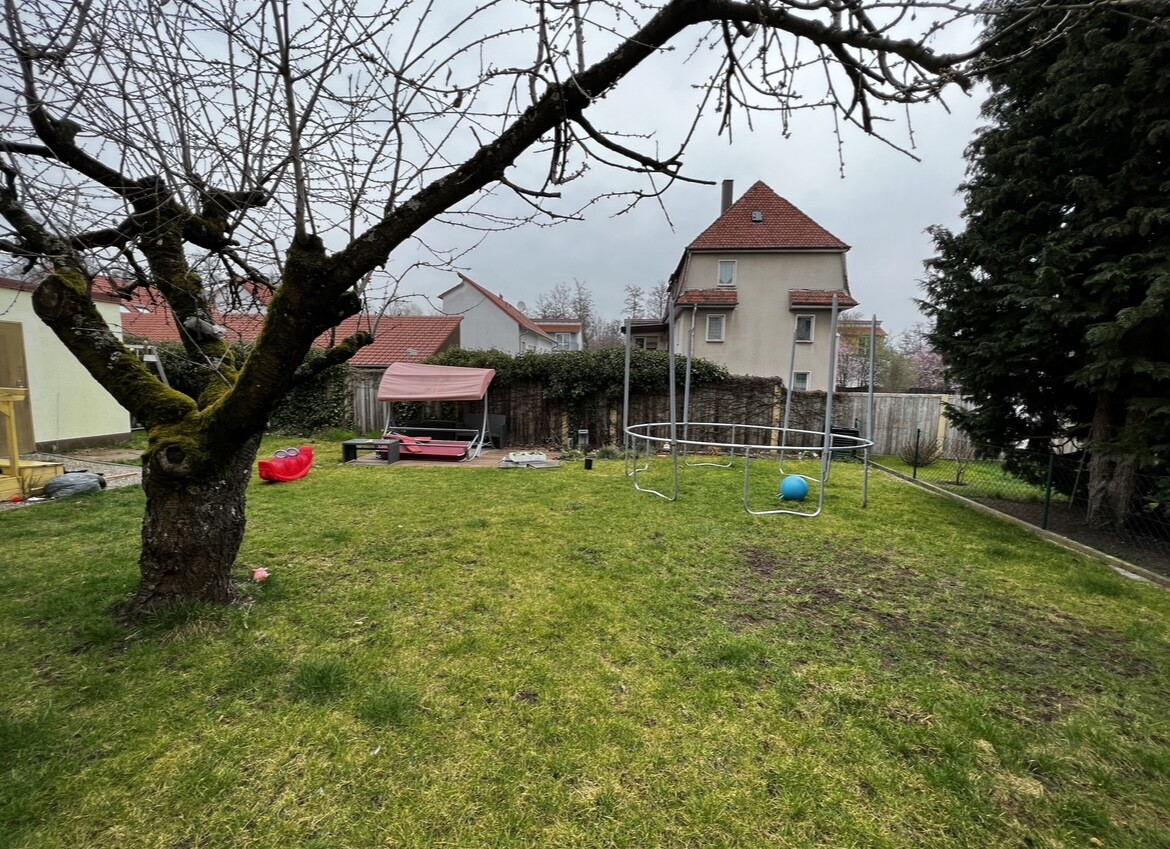 Gartenansicht: Großer Baum links, Trampolin, Pavillon und Spielzeug, Häuser im Hintergrund