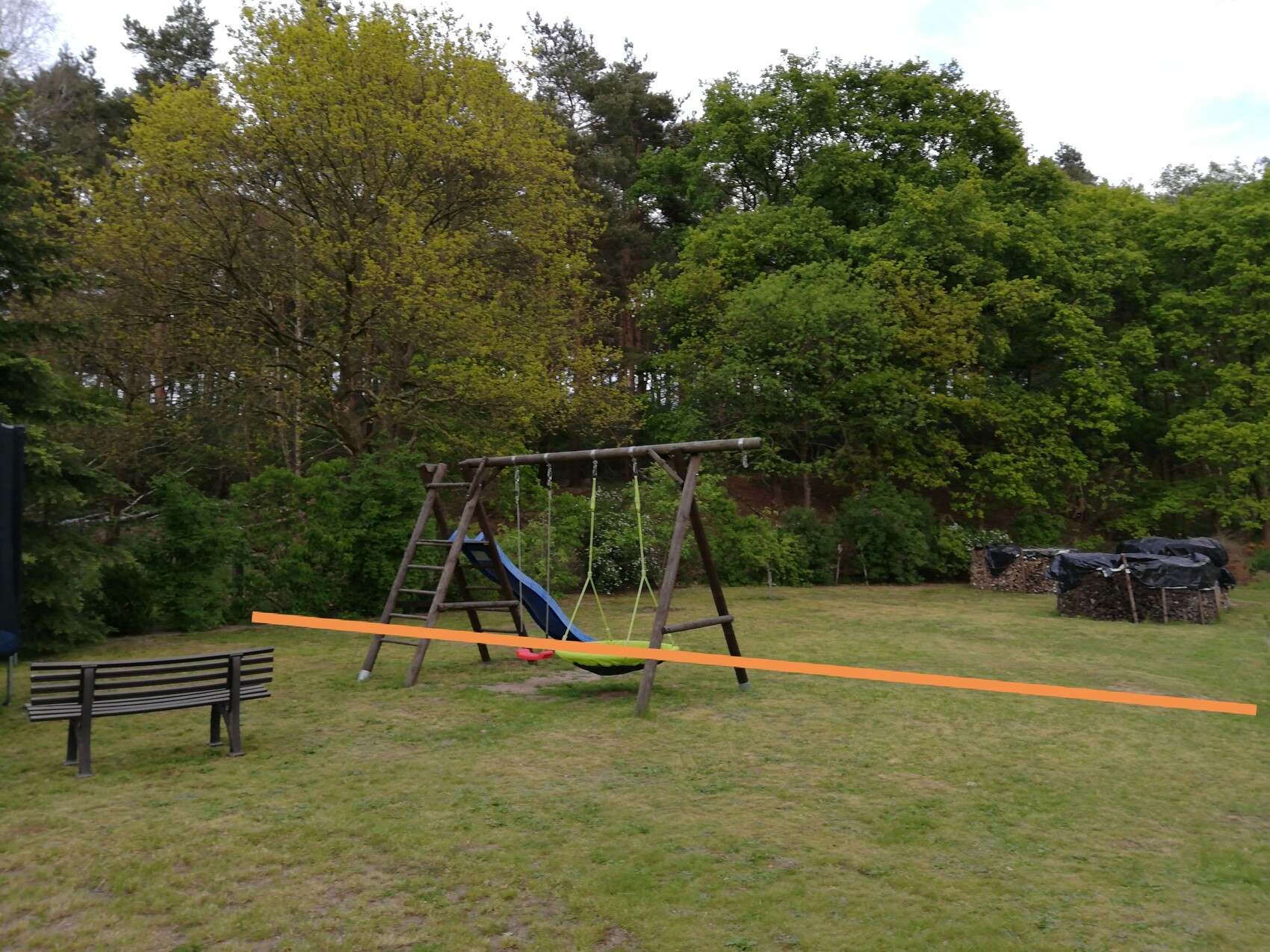 Spielplatz im Garten: Schaukelgestell mit blauer Rutsche, orange Absperrband, Bank links, Holzstapel