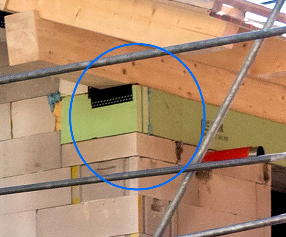 Baustelle: Mauerwerk, grüne Dämmplatten, schwarzes Lüftungsgitter in der Ecke, Gerüststangen.