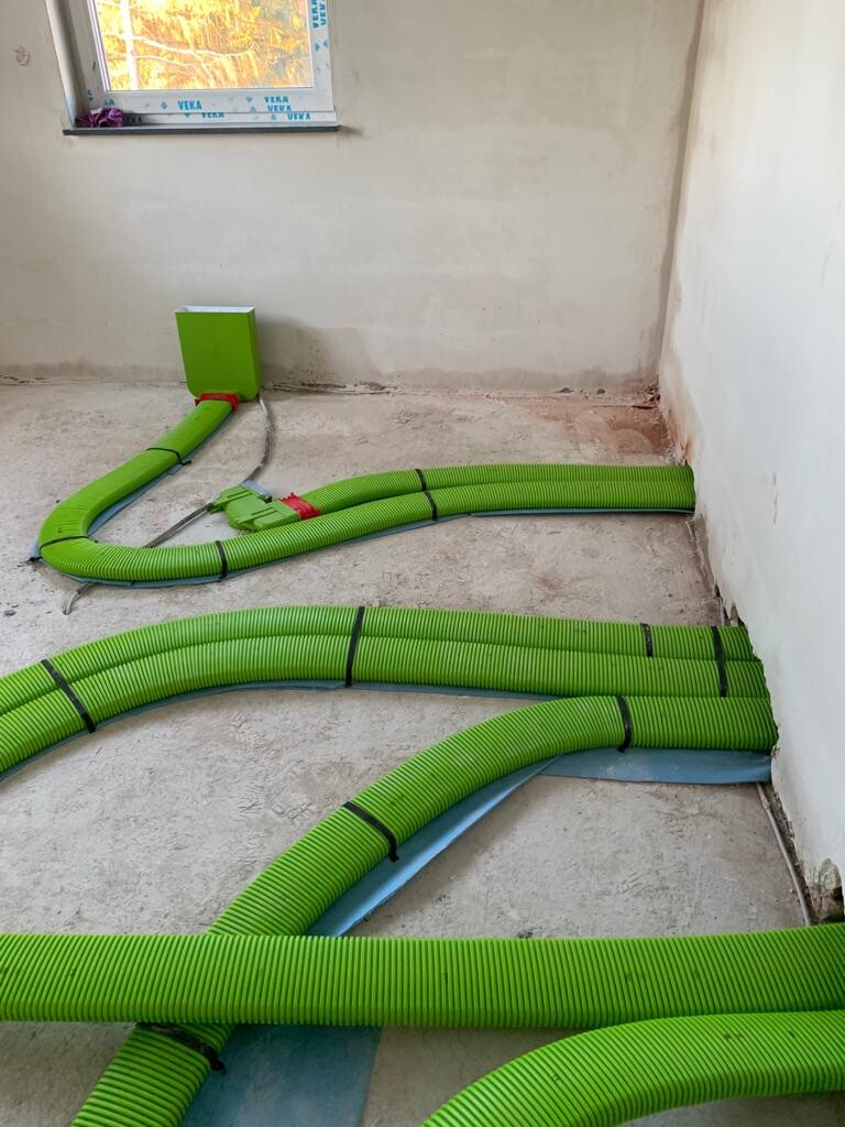 Grüne flexible Lüftungsrohre verlaufen über Betonboden zu einer Wandbox im Neubau.