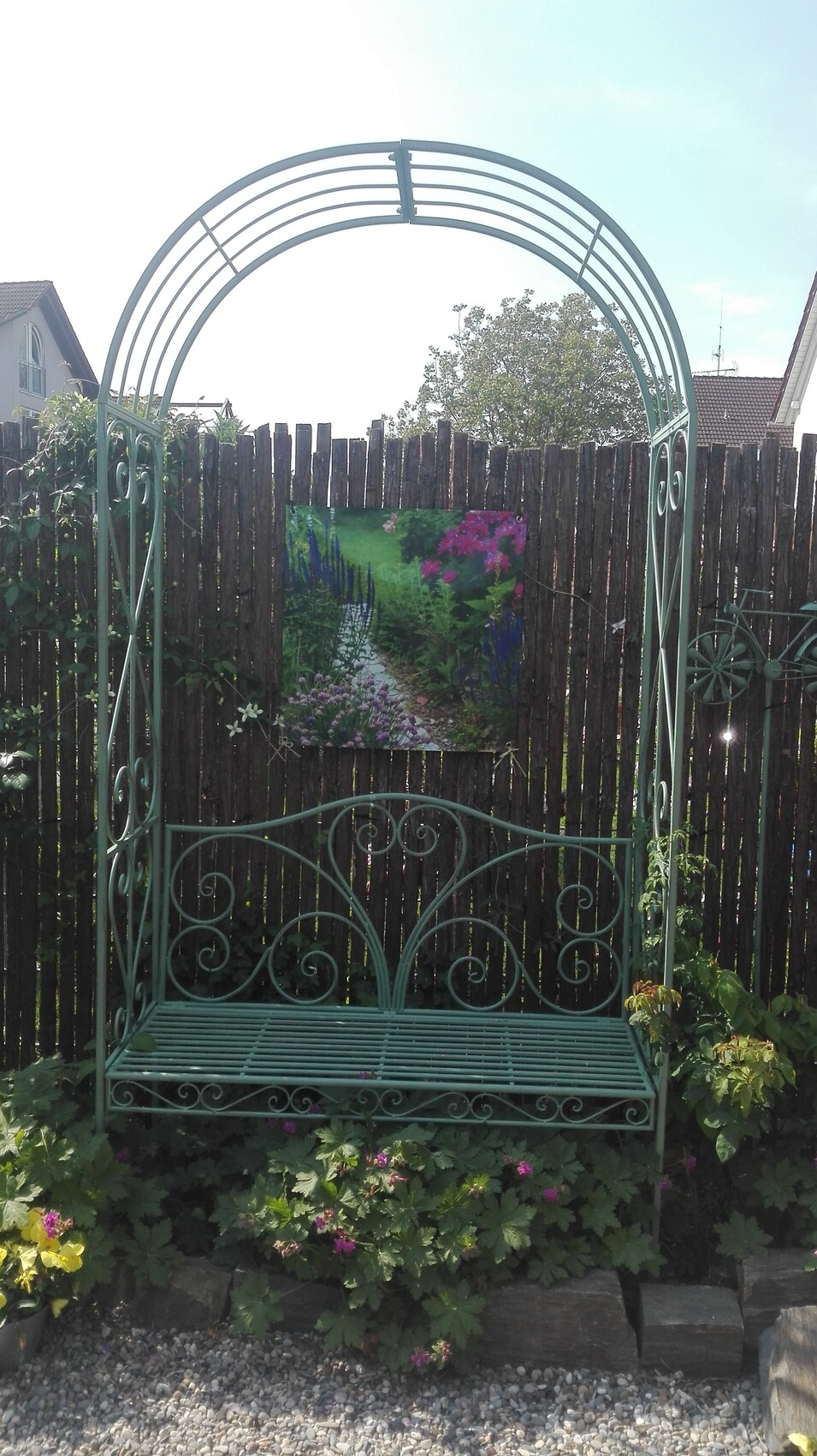 Grüne dekorative Metallbogen-Gartenbank vor Holzzaun, umgeben von Blumen; Gartenbild im Hintergrund.