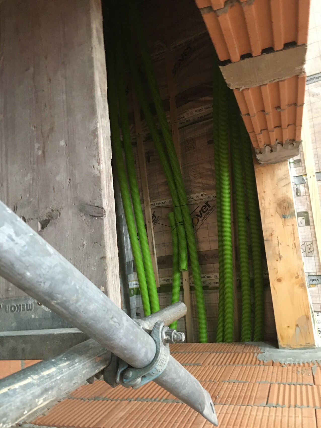 Mehrere grüne Kabelrohre führen an einer Wand neben Holzrahmen und Gerüststangen vorbei.