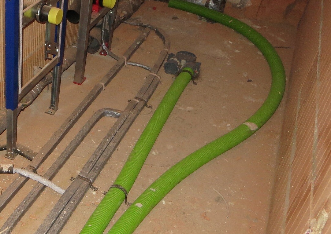 Grüne flexible Rohre liegen auf staubigem Boden neben Metallrohren in einem Bau-/Installationsraum.