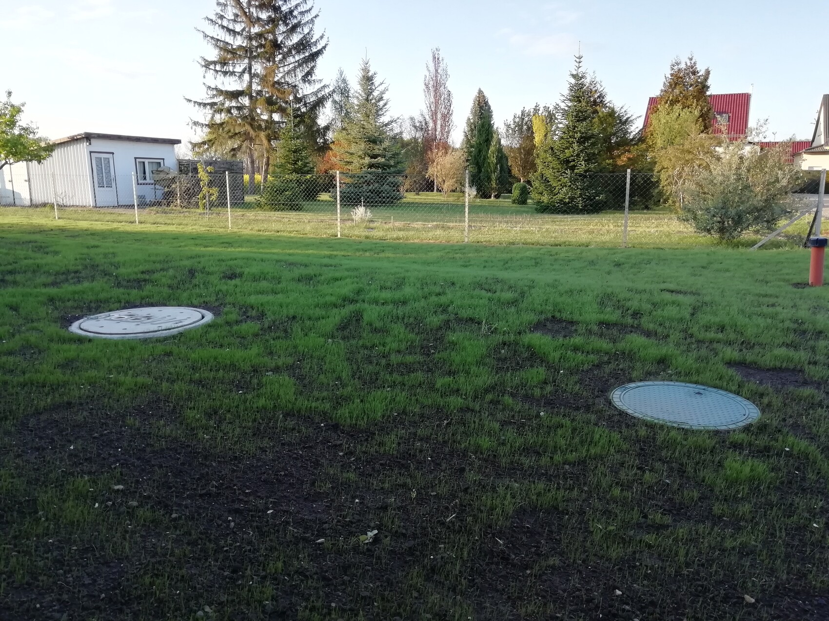 Grüner Rasen mit zwei runden Metalldeckeln; Zaun und Häuser im Hintergrund.